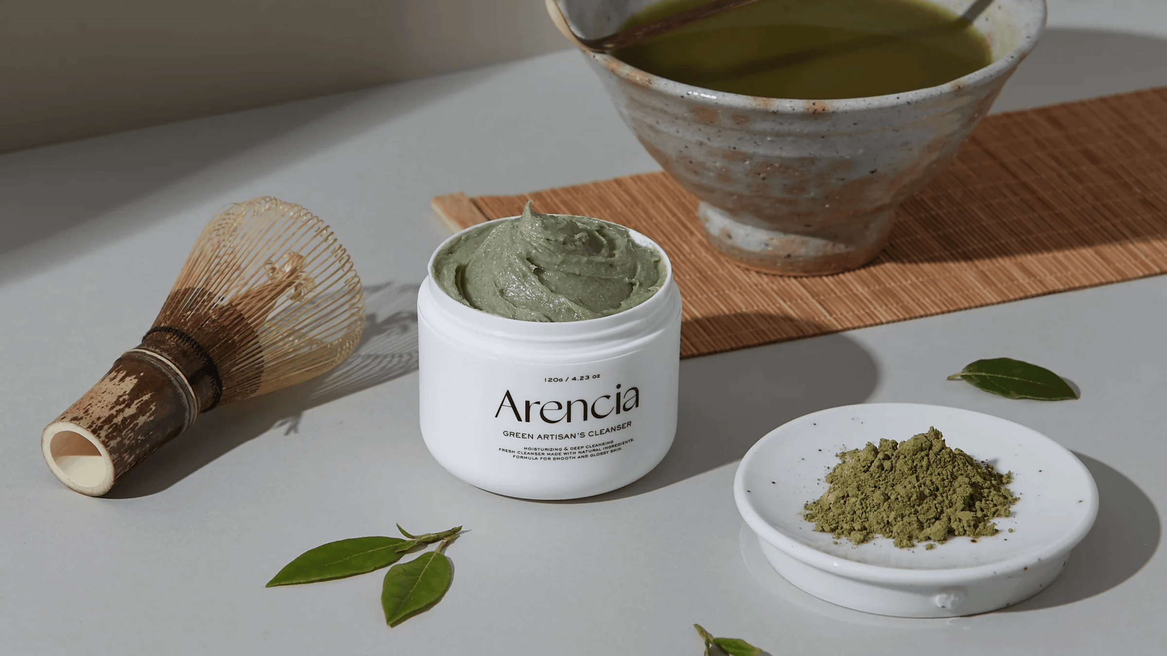 Arencia Green Artisan's Cleanser i en vit burk med grön rengöringskräm, omgiven av teblad, bambuvisp, matchapulver och en keramikskål med grönt te på en bambumatta.