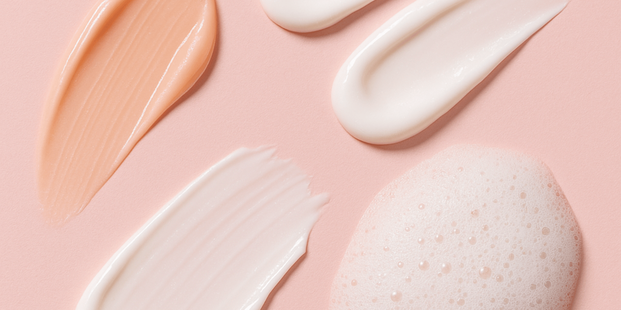 Flera typer av hudvårdsprodukter applicerade på en rosa bakgrund – inkluderar en persikofärgad gel, två olika vita krämer och ett mjukt skum, som visar upp olika texturer och konsistenser.