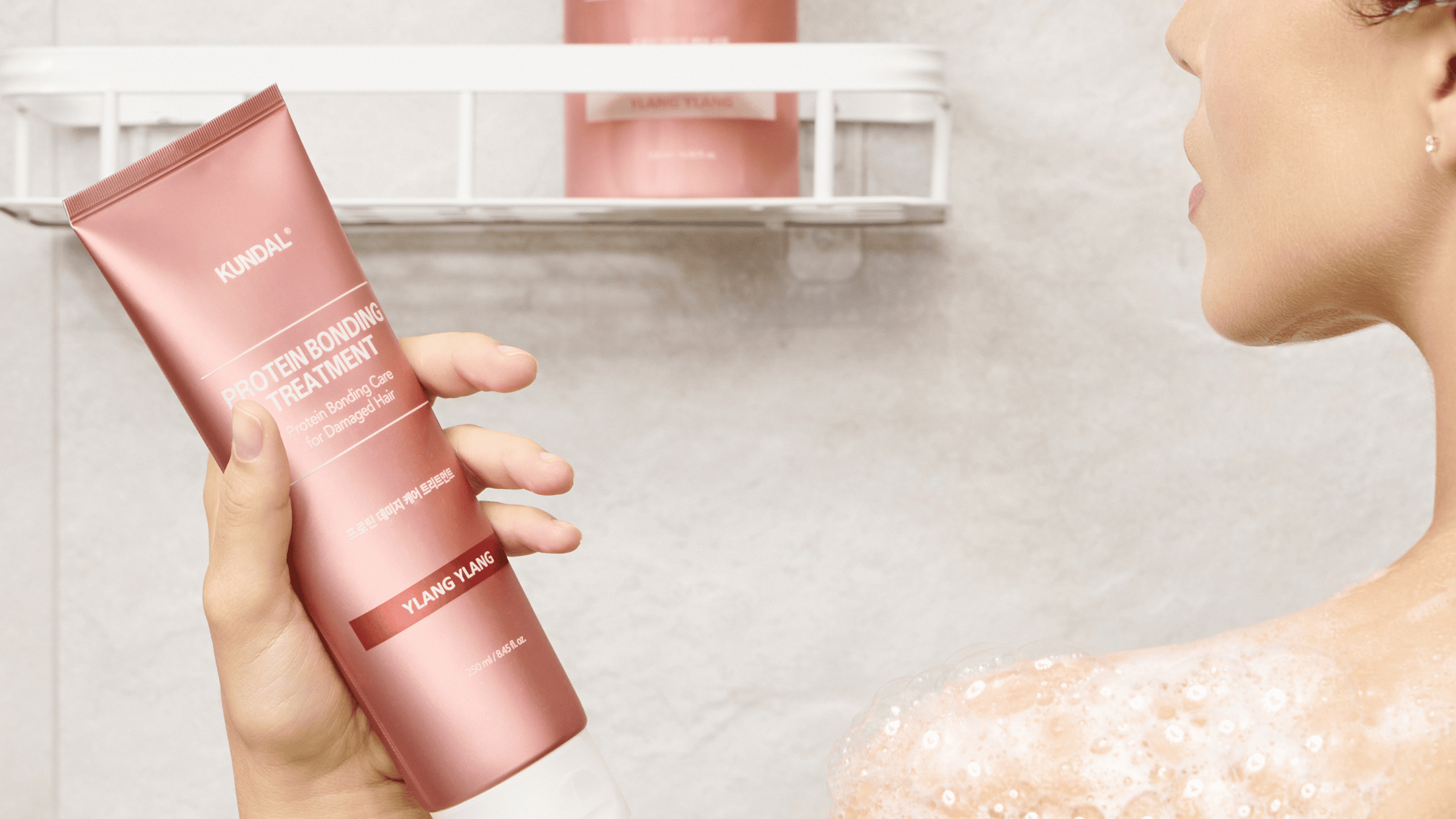 Person i duschen håller KUNDAL Protein Bonding Treatment Ylang Ylang, en hårbehandling för skadat hår, med lödder på huden och matchande flaska i hyllan.