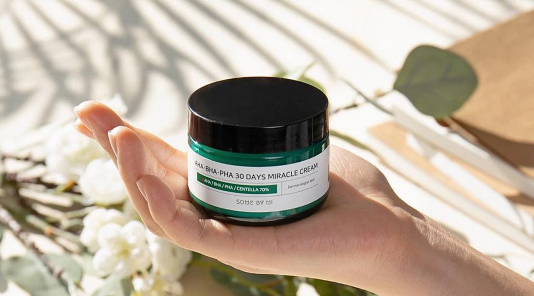 En hand håller i en grön burk med svart lock – AHA BHA PHA 30 Days Miracle Cream från SOME BY MI – mot en bakgrund med blommor och skuggor från solljus.