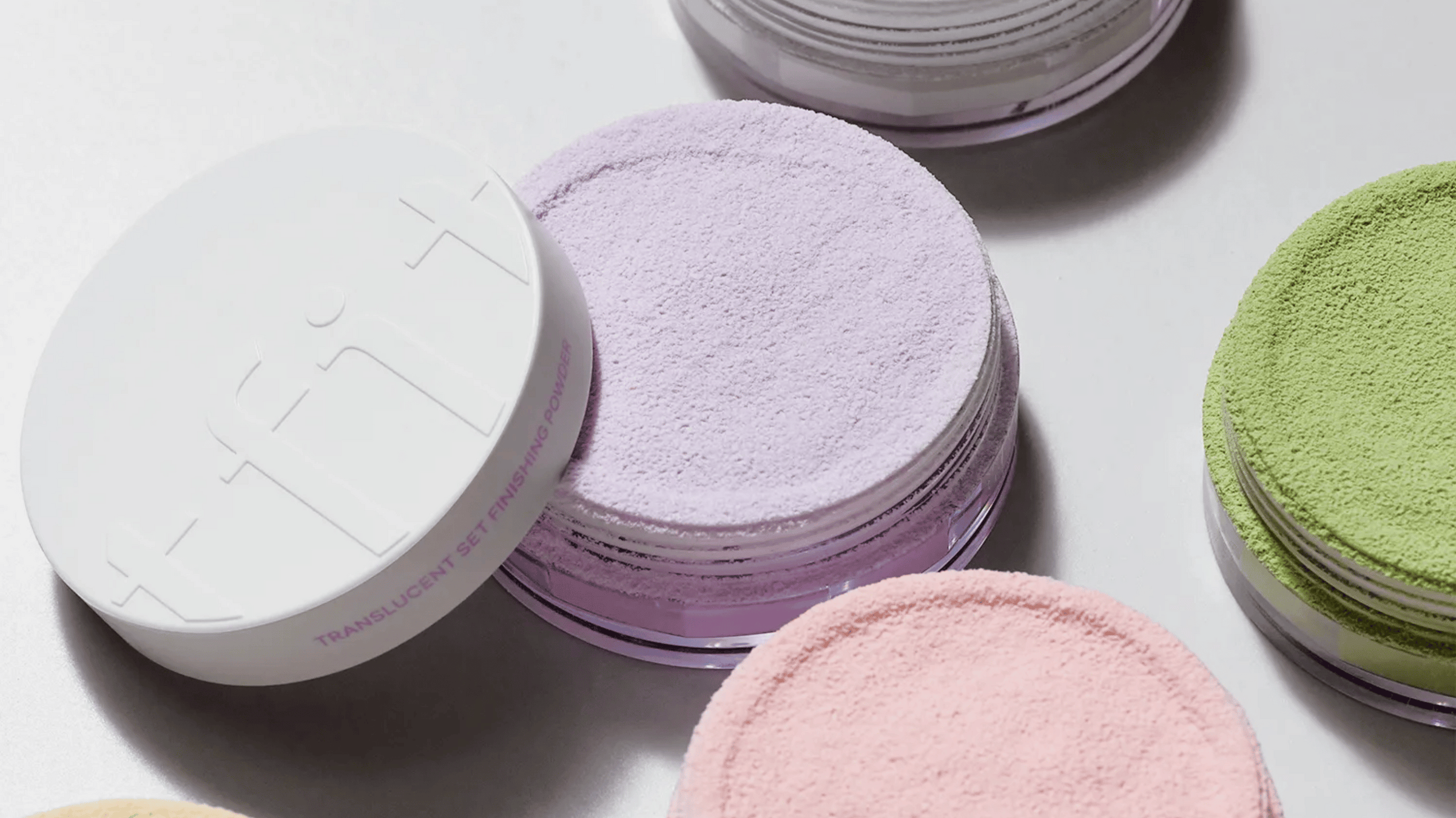 Närbild på olika nyanser av lösa, genomskinliga setting-puder i behållare. Pudren visas i lavendel, rosa och gröna nyanser, där lavendelpudret är i förgrunden.