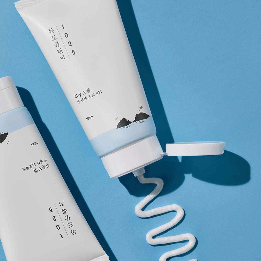 Dve tuby 1025 Dokdo Cleanser na modrom pozadí, jedna s otvoreným uzáverom a druhá s vlnitou čiarou vytlačeného čistiaceho prostriedku.