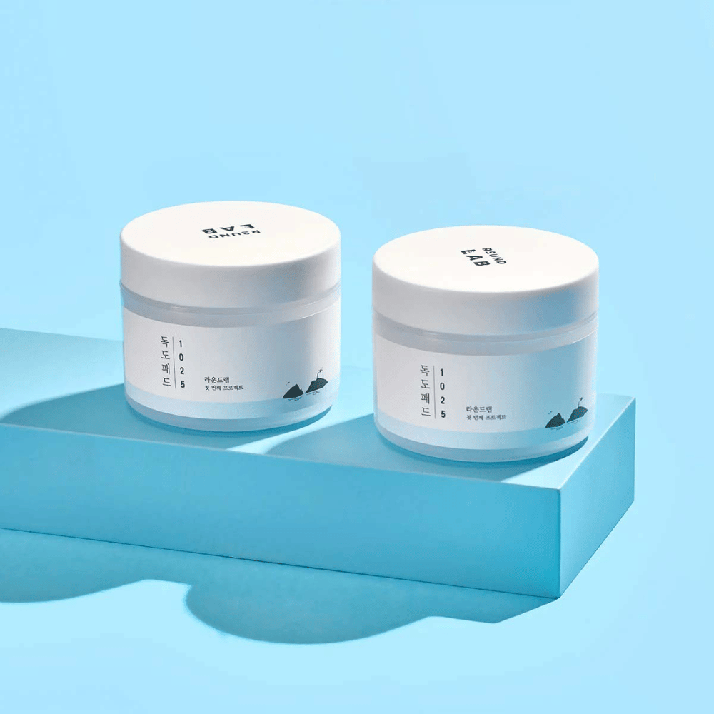 Dve biele nádoby 1025 Dokdo Pads, umiestnené na svetlomodrej polici, ktorá vrhá odraz, na modrom pozadí.