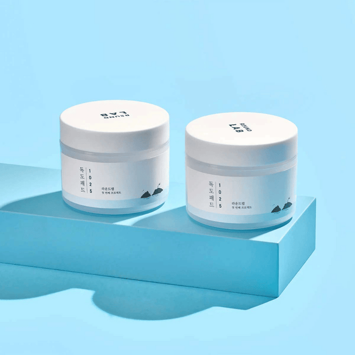 Dve biele nádoby 1025 Dokdo Pads, umiestnené na svetlomodrej polici, ktorá vrhá odraz, na modrom pozadí.