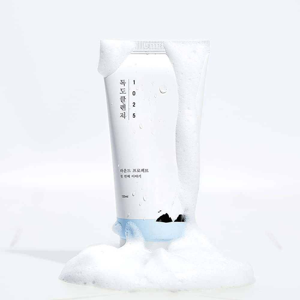 1025 Dokdo Cleanser v bielej tube pokrytej penou, s kvapkami vody a penou okolo dna, na svetlom pozadí.