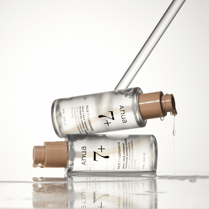 Obrázok zobrazuje dve priehľadné fľaše "Anua 77 Rice + Ceramide Hydrating Barrier Serum" s kvapkami stekajúcimi z pipety. Sérum, ktoré je obohatené o ryžu a ceramidy, je určené na hydratáciu a posilnenie kožnej bariéry. Fľaše majú elegantný dizajn, ktorý posilňuje prémiový pocit z produktu.