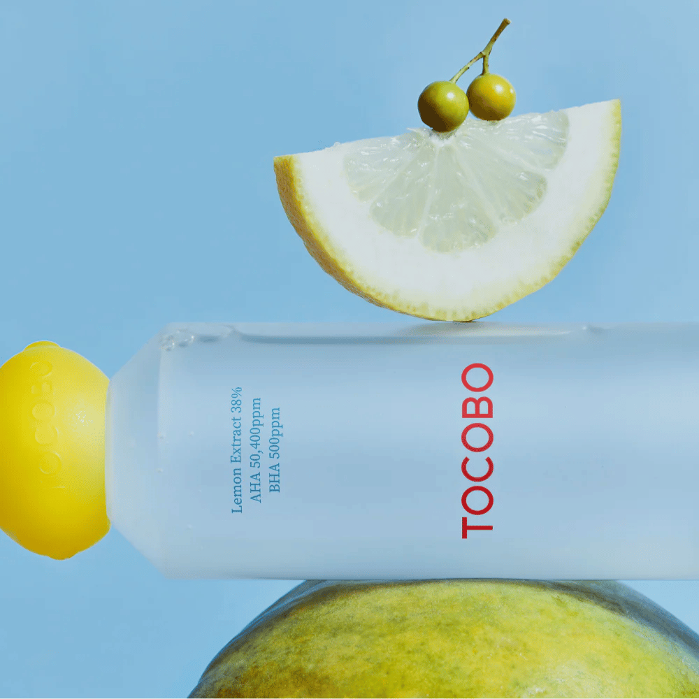 Fľaša s TOCOBO AHA BHA Lemon Toner, zdobená uzáverom v tvare citróna. Plátok citróna a malé zelené bobule balansujú na fľaši proti modrému pozadiu.