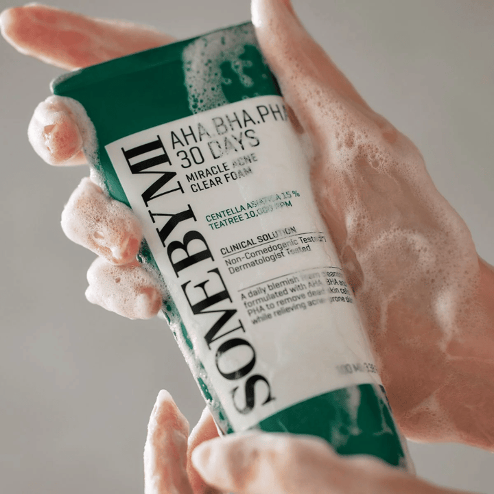 Osoba drží tubu "SOME BY MI AHA, BHA, PHA 30 Days Miracle Acne Clear Foam" s penivým obsahom na rukách. Produkt je určený na každodenné použitie na boj proti akné a čistenie pórov s jeho aktívnymi zložkami ako centella, tea tree olej a kyseliny ako AHA, BHA a PHA.