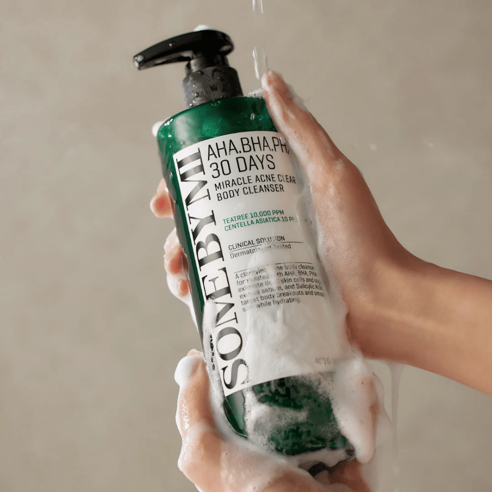 Osoba drží fľašu SOME BY MI AHA.BHA.PHA 30 Days Miracle Acne Clear Body Cleanser. Fľaša je zelená s bielou etiketou a je pokrytá penou z čistiaceho prostriedku. Osoba sa umýva produktom, zatiaľ čo voda steká po rukách.