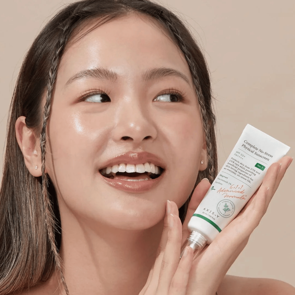 Obrázok zobrazuje mladú ženu, ktorá sa šťastne usmieva a drží tubu s AXIS-Y Complete No-Stress Physical Sunscreen. Má žiarivý a prirodzený vzhľad s minimálnym líčením, čo zvýrazňuje produkt, ktorý predstavuje.