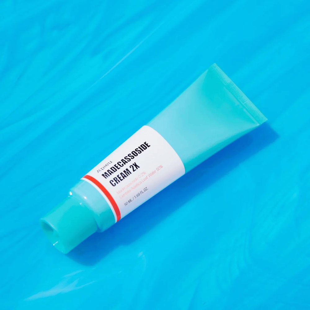 Tuba A'PIEU Madecassoside Cream 2X leží diagonálne cez modré pozadie s vráskami pripomínajúcimi vodu. Tuba má biele telo so svetlým tyrkysovým uzáverom a červeno-oranžovú etiketu, na ktorej je jasne napísaný názov produktu a aktívna zložka Madecassoside 2%. Na etikete je tiež uvedené množstvo, ktoré je 50 ml / 1.8 fl oz. Svetlá a farebná prezentácia dodáva čerstvý a energický pocit.