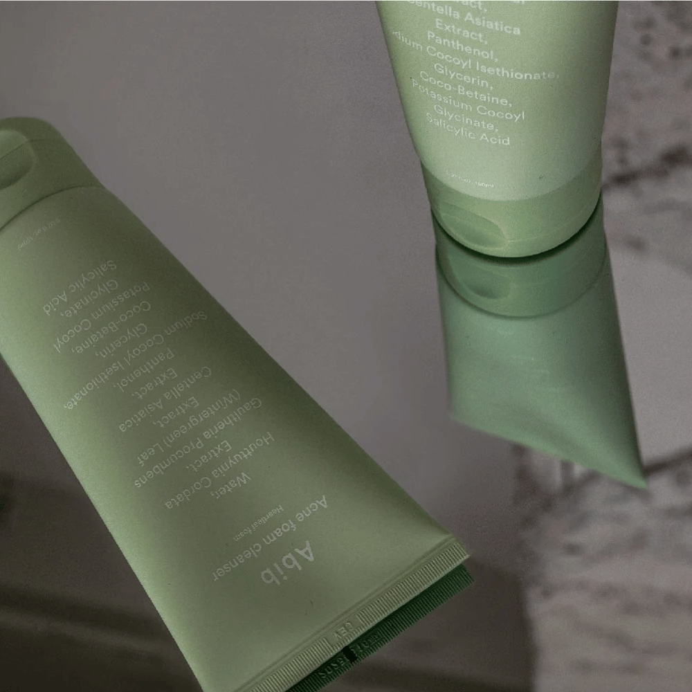Zelená tuba Abib Acne Foam Cleanser Heartleaf ležiaca na zrkadlovom stole s prednou stranou a textom produktu odrazeným. Ingrediencie ako kyselina salicylová, centella a extrakt z listov srdca sú jasne viditeľné na obale.