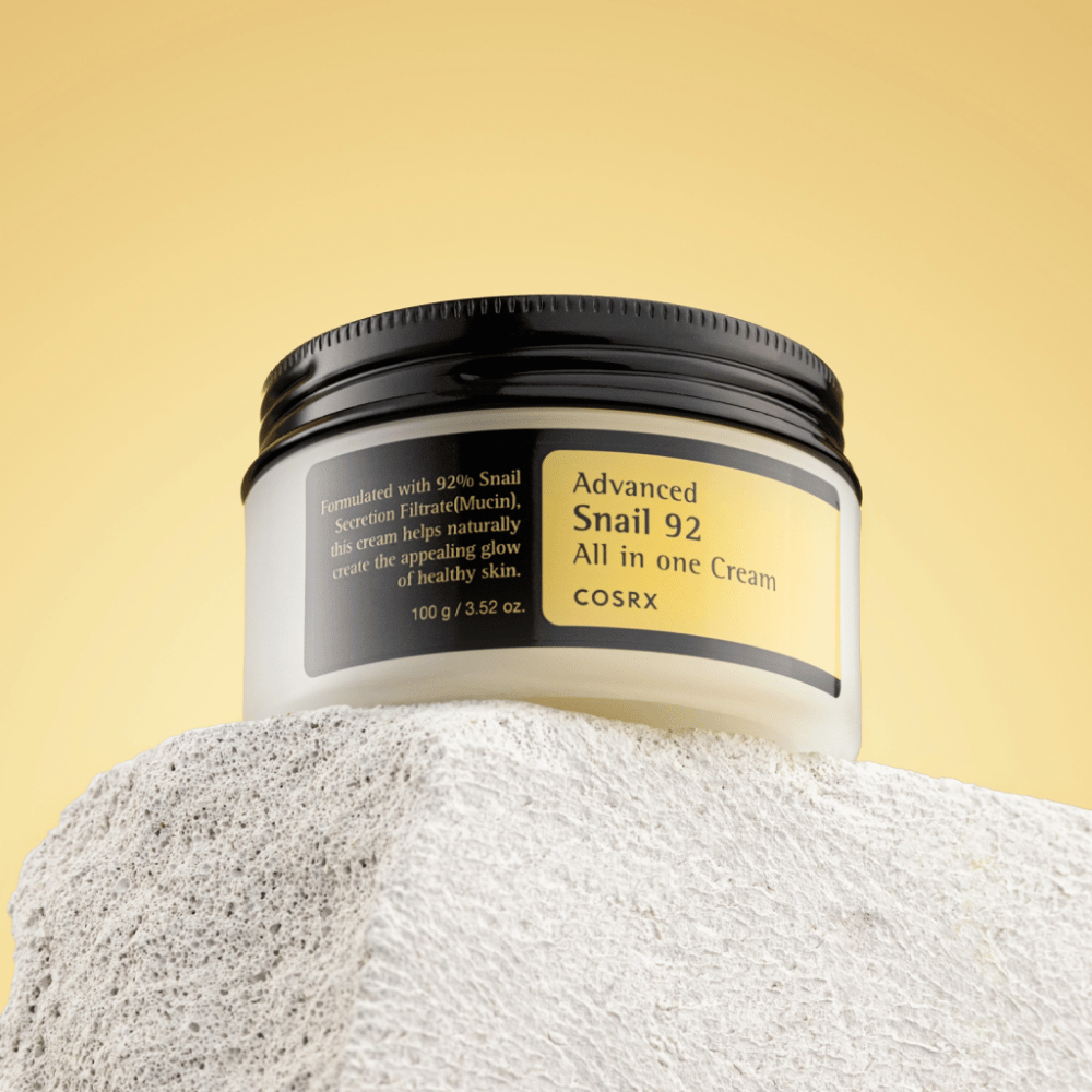 Nádoba COSRX Advanced Snail 92 All in one Cream umiestnená na kamenistom povrchu so žltým pozadím.