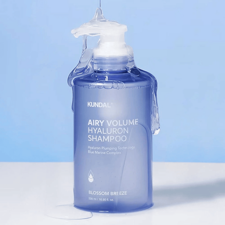KUNDAL Airy Volume Hyaluron Shampoo Blossom Breeze v modrej pumpovacej fľaši, s kyselinou hyalurónovou a Blue Marine Complex, fotografovaný s tečúcou vodou.