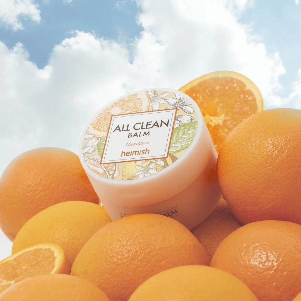 Pohár Heimish All Clean Balm Mandarin umiestnený na kope pomarančov, s modrou oblohou v pozadí.