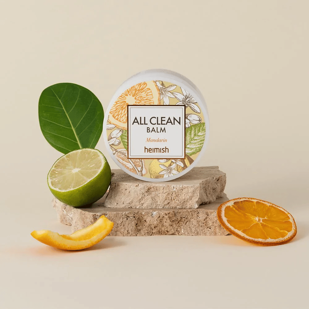 Pohár Heimish All Clean Balm Mandarin umiestnený na kameňoch, obklopený mandarínkovými šupkami a citrusovými listami, pred svetlým pozadím.