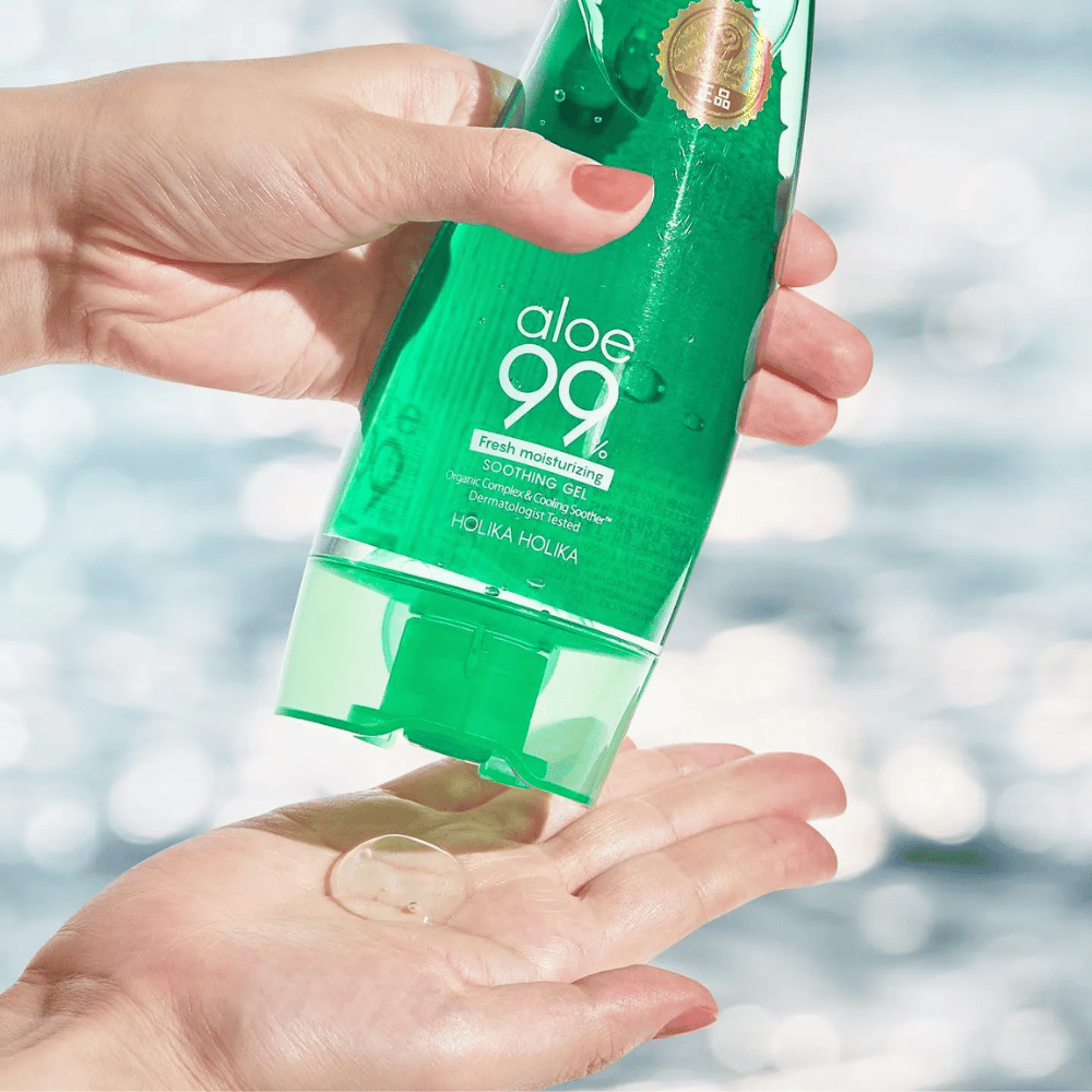 Ruka drží zelenú tubu s Holika Holika Aloe 99% Soothing Gel, zatiaľ čo druhá ruka zachytáva kvapku gélu, ktorý sa dávkuje. Pozadie je rozmazané a zobrazuje vodnatý, trblietavý vzor.