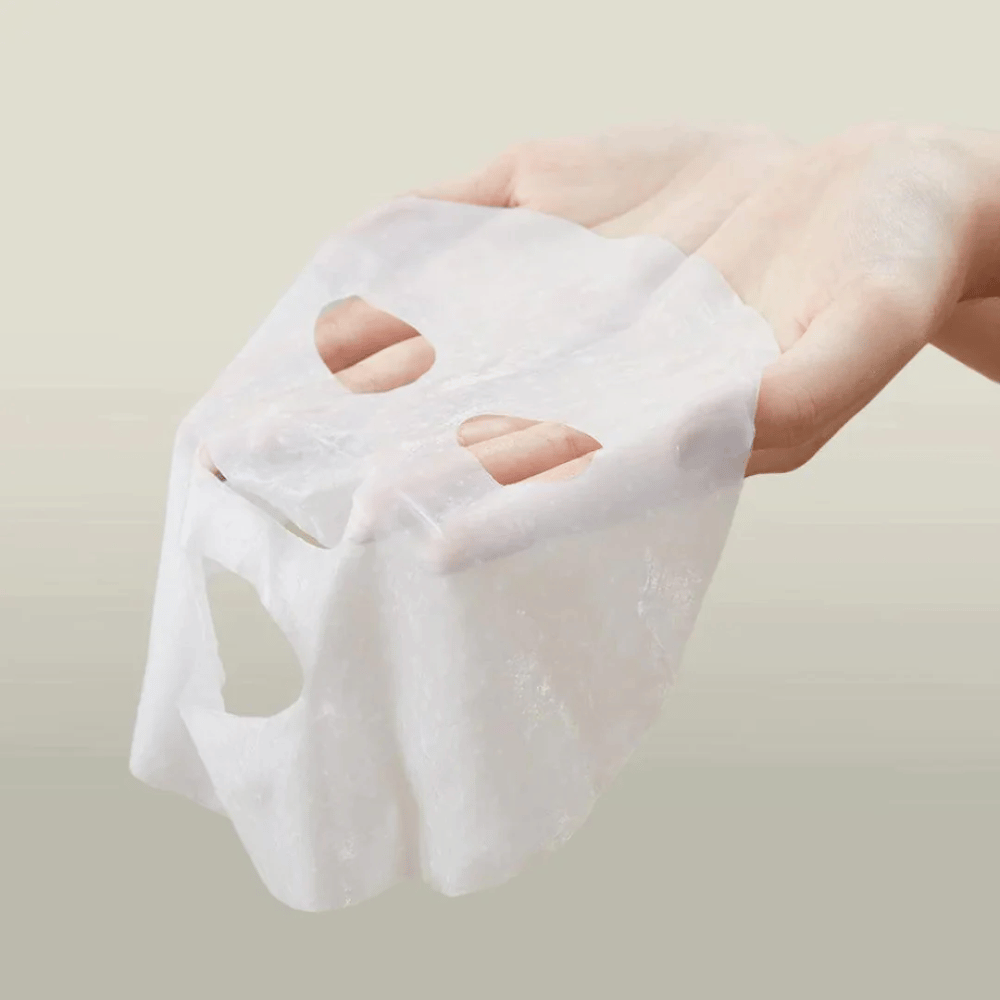 Obrázok ukazuje ruku držiacu pleťovú masku značky "Anua". Zdá sa, že ide o "Heartleaf Cream Mask Night Solution", čo naznačuje, že ide o nočnú masku na starostlivosť o pleť s extraktom z listov srdca. Maska vyzerá byť vyrobená z látky a je nasiaknutá krémom alebo sérom, čo je typické pre kórejské produkty na starostlivosť o pleť, ktoré sa zameriavajú na intenzívnu hydratáciu a starostlivosť o pleť počas noci.