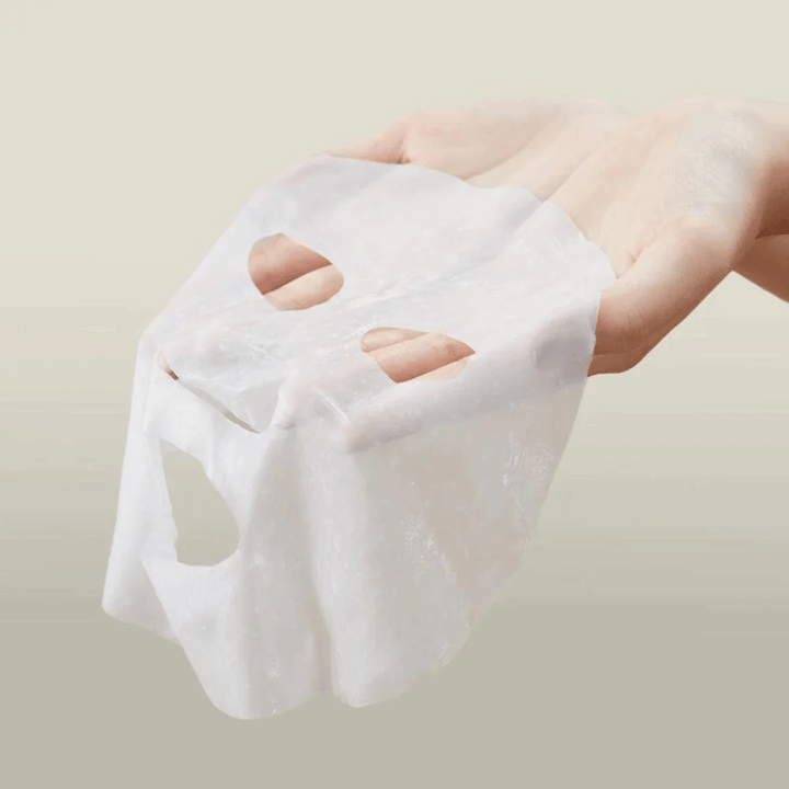 Obrázok ukazuje ruku držiacu pleťovú masku značky "Anua". Zdá sa, že ide o "Heartleaf Cream Mask Night Solution", čo naznačuje, že ide o nočnú masku na starostlivosť o pleť s extraktom z listov srdca. Maska vyzerá byť vyrobená z látky a je nasiaknutá krémom alebo sérom, čo je typické pre kórejské produkty na starostlivosť o pleť, ktoré sa zameriavajú na intenzívnu hydratáciu a starostlivosť o pleť počas noci.