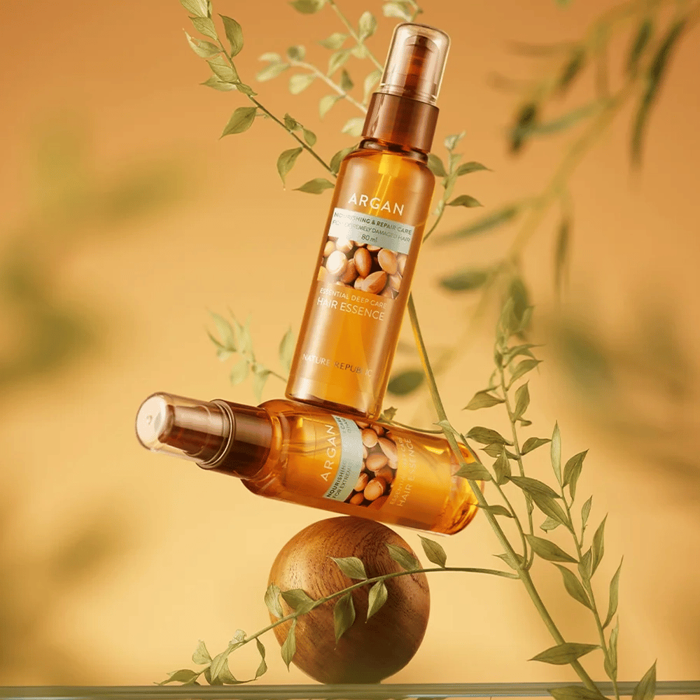 Dve fľaše Nature Republic Argan Essential Moisture Hair Oil umiestnené medzi zelenými listami – hodvábny lesk pre suché vlasy.