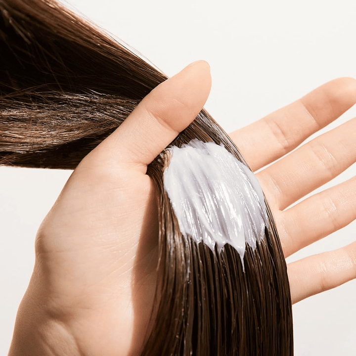 Ruka aplikuje Nature Republic Argan Essential Moisture Repairing Hair Mask na mokré vlasy – hydratačná maska, ktorá posilňuje a zjemňuje vlasy.