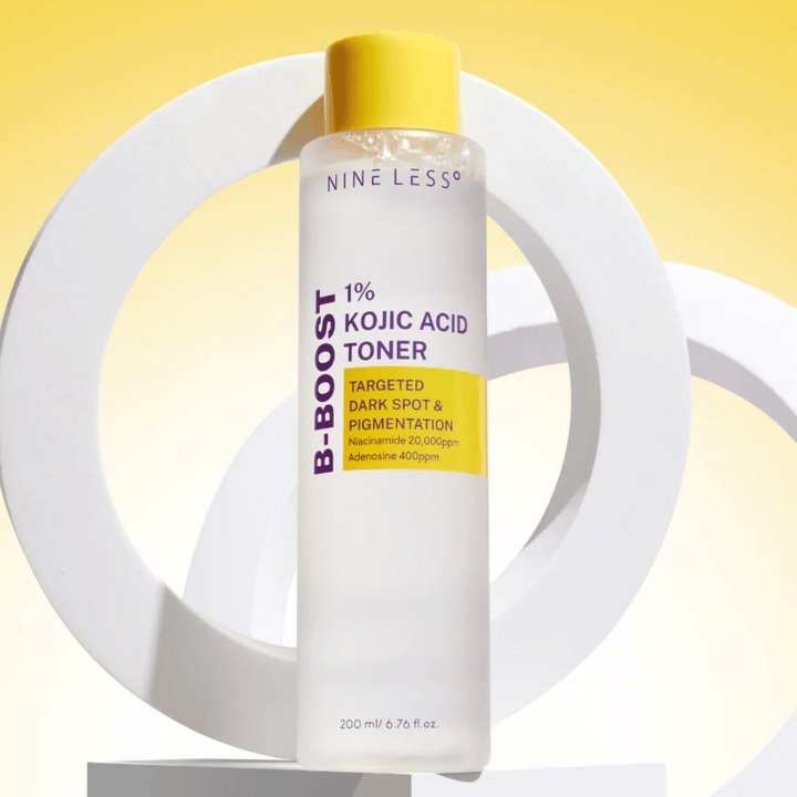 NINE LESS B-Boost 1% Kojic Acid Toner stojaci na žltom pozadí s textom "Cielené tmavé škvrny a pigmentácia". Toner na vyrovnávanie tónu pleti s niacínamidom a adenozínom.