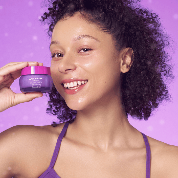 Žena so žiarivou pleťou drží EQQUALBERRY Bakuchiol Plumping Capsule Cream a usmieva sa pred fialovým pozadím.
