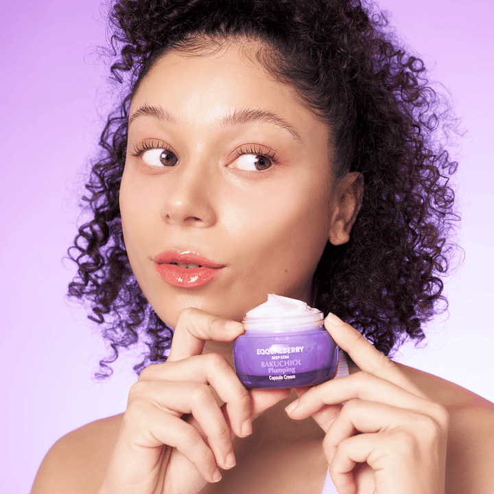 Žena drží EQQUALBERRY Bakuchiol Plumping Capsule Cream v ruke a ukazuje svoju hydratovanú, žiarivú pleť na fialovom pozadí.