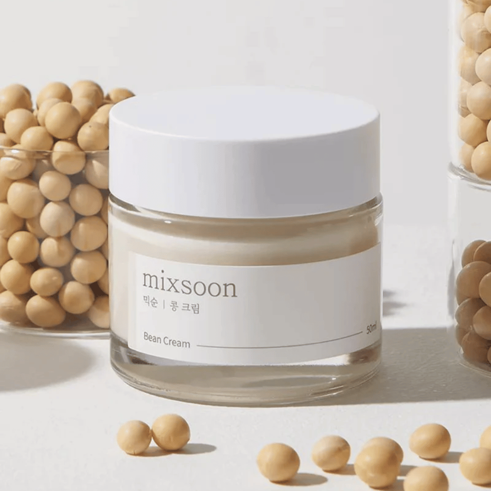 Priehľadný pohár s bielou etiketou a viečkom, označený "mixsoon 팥 크림" a "Bean Cream" s uvedeným objemom "50ml". Pohár obsahuje svetložltý krém a je umiestnený na svetlom povrchu. Vedľa pohára je sklenená nádoba plná svetlohnedých fazúľ, ktoré sú rozliate po povrchu, čo naznačuje, že krém obsahuje fazuľový extrakt. Pozadie a osvetlenie vytvárajú teplú a prirodzenú atmosféru.