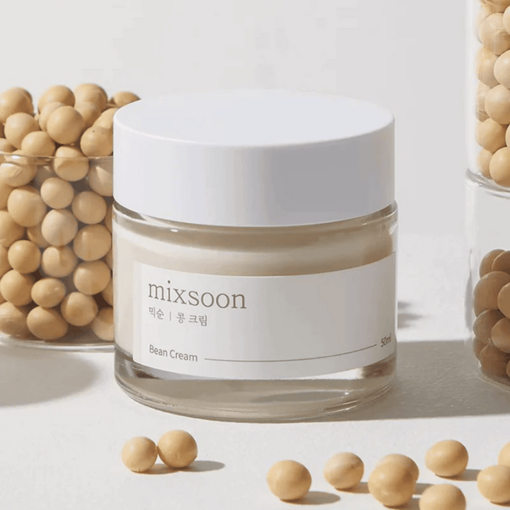 Priehľadný pohár s bielou etiketou a viečkom, označený "mixsoon 팥 크림" a "Bean Cream" s uvedeným objemom "50ml". Pohár obsahuje svetložltý krém a je umiestnený na svetlom povrchu. Vedľa pohára je sklenená nádoba plná svetlohnedých fazúľ, ktoré sú rozliate po povrchu, čo naznačuje, že krém obsahuje fazuľový extrakt. Pozadie a osvetlenie vytvárajú teplú a prirodzenú atmosféru.