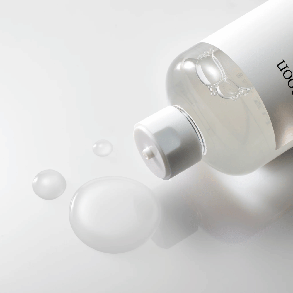 Priehľadná fľaša leží na boku na lesklom povrchu s ľahkým tonerom, ktorý pomaly vyteká a tvorí kvapky a väčšiu bublinu na povrchu. Fľaša má bielu etiketu a biely pumpovací mechanizmus. Osvetlenie je jemné a vytvára čistú a upokojujúcu atmosféru, zatiaľ čo ľahká konzistencia a jasnosť tonera sú zdôraznené tým, ako sa šíri po povrchu.