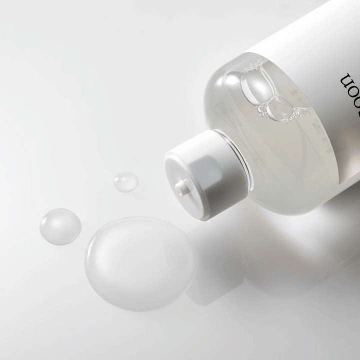 Priehľadná fľaša leží na boku na lesklom povrchu s ľahkým tonerom, ktorý pomaly vyteká a tvorí kvapky a väčšiu bublinu na povrchu. Fľaša má bielu etiketu a biely pumpovací mechanizmus. Osvetlenie je jemné a vytvára čistú a upokojujúcu atmosféru, zatiaľ čo ľahká konzistencia a jasnosť tonera sú zdôraznené tým, ako sa šíri po povrchu.