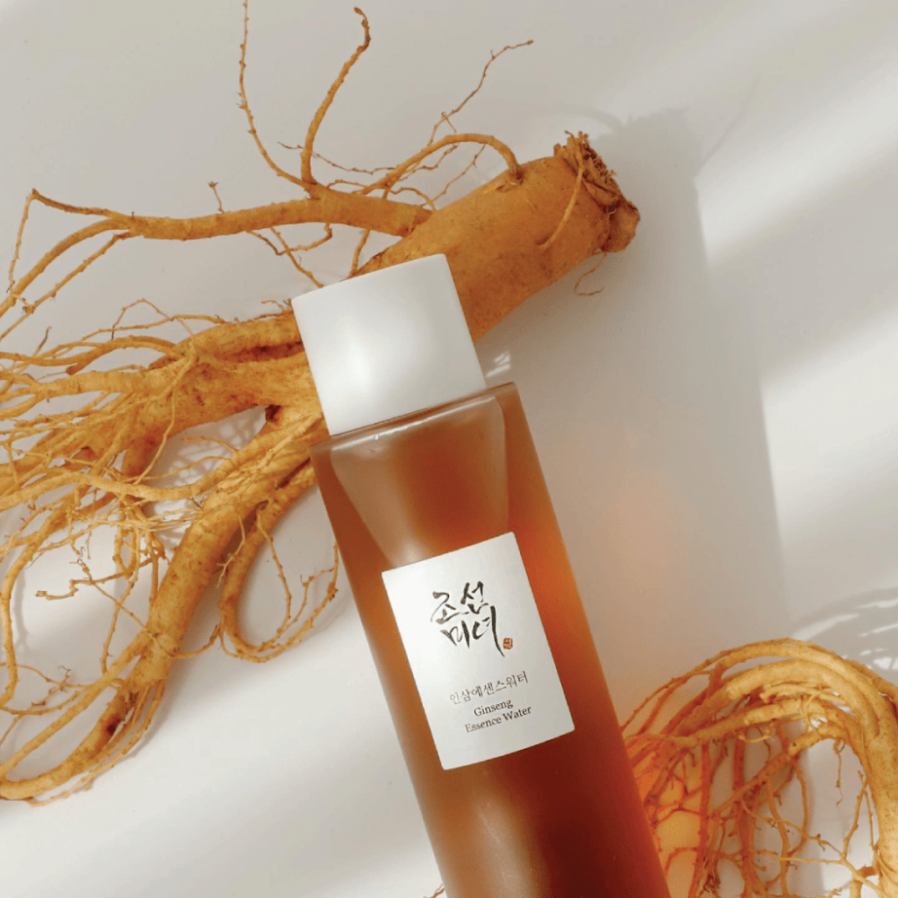 Hnedá fľaša Beauty of Joseon Ginseng Essence Water s bielym uzáverom leží vedľa čerstvého koreňa ženšenu na bielom pozadí.