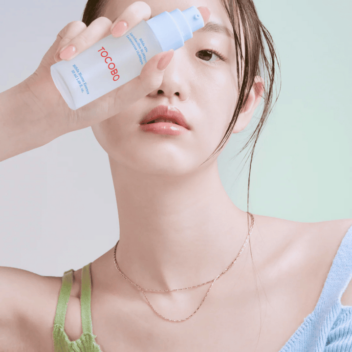 Mladá žena s jemným make-upom a prirodzeným tónom pleti drží fľašu TOCOBO Bifida Biome Essence pred tvárou. Má na sebe svetlomodré tričko so zelenými ramienkami a tenký zlatý náhrdelník na svetlom pozadí.