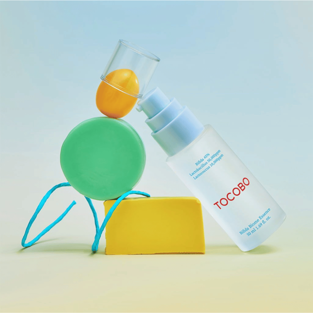 TOCOBO Bifida Biome Essence v matnej pumpovacej fľaši s červeným logom, umiestnená na farebnom pozadí so zelenými, žltými a oranžovými geometrickými tvarmi. Text na fľaši uvádza Bifida 5 %, Lactobacillus 3 000 ppm a Lactococcus 1 000 ppm.