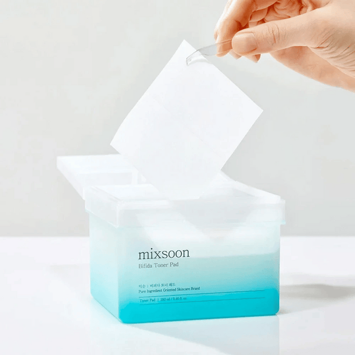 Priehľadná nádoba s modro-bielym prechodom obsahujúca Bifida tonerové podložky. Ruka vyberá tonerovú podložku z nádoby, ktorá je označená "mixsoon Bifida Toner Pad" a obsahuje 280 ml (9,46 fl. oz.) tonerových podložiek určených pre starostlivosť o pleť.