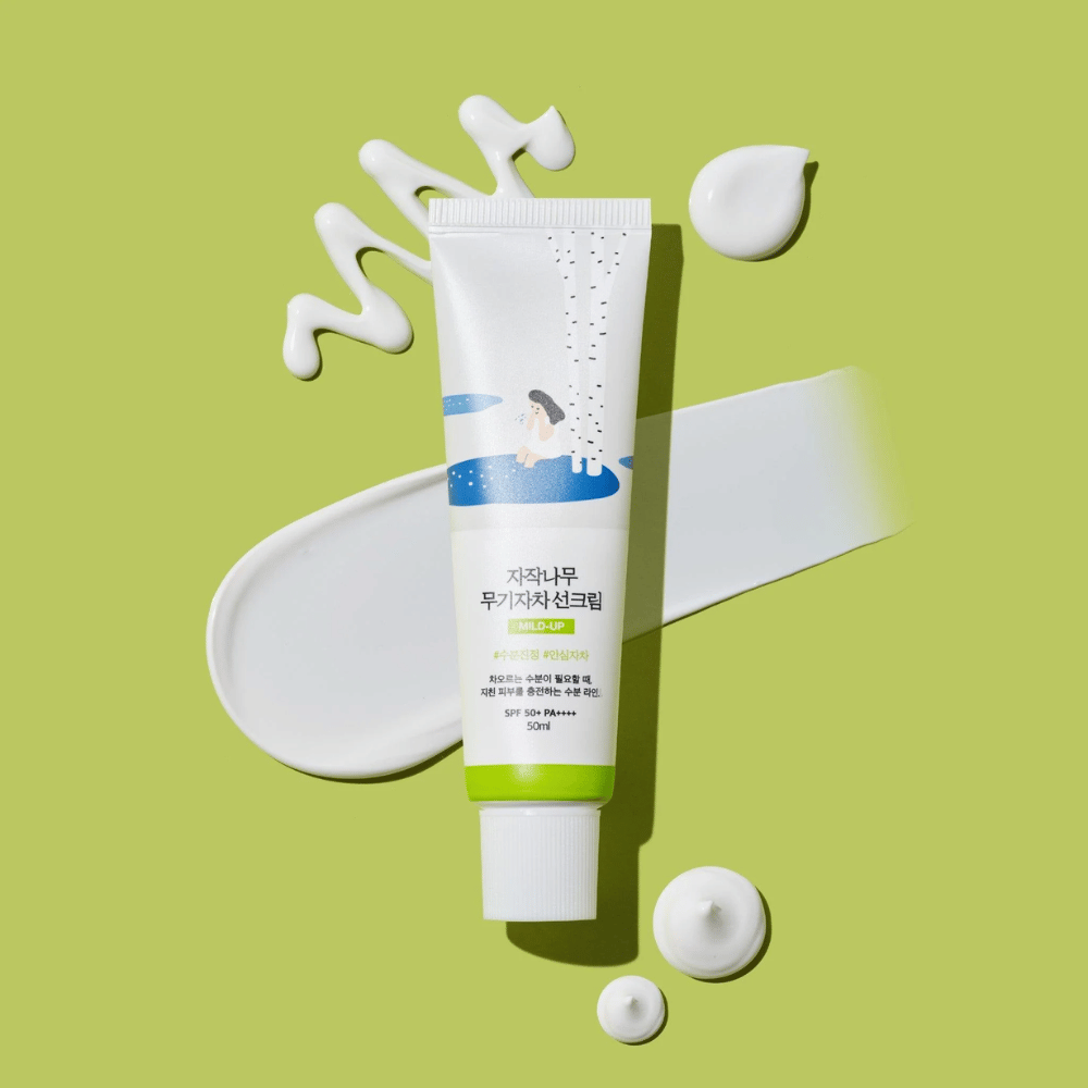 Birch Juice Moisturizing Mild Up Sunscreen – biela tuba so zeleným základom na zelenej pozadí, obklopená rozmazaným opaľovacím krémom a kvapkami, SPF 50+ PA++++, 50 ml.