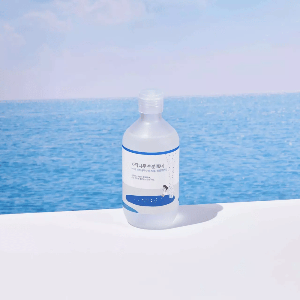 Fľaša ROUND LAB Birch Juice Moisturizing Toner stojí na bielom povrchu s morom a modrou oblohou v pozadí