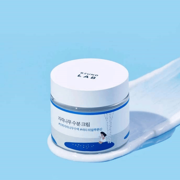 Round Lab Birch Juice Moisturizing Cream v modrastom obale s bielym viečkom, stojaci v vrstve krému na modrom pozadí.