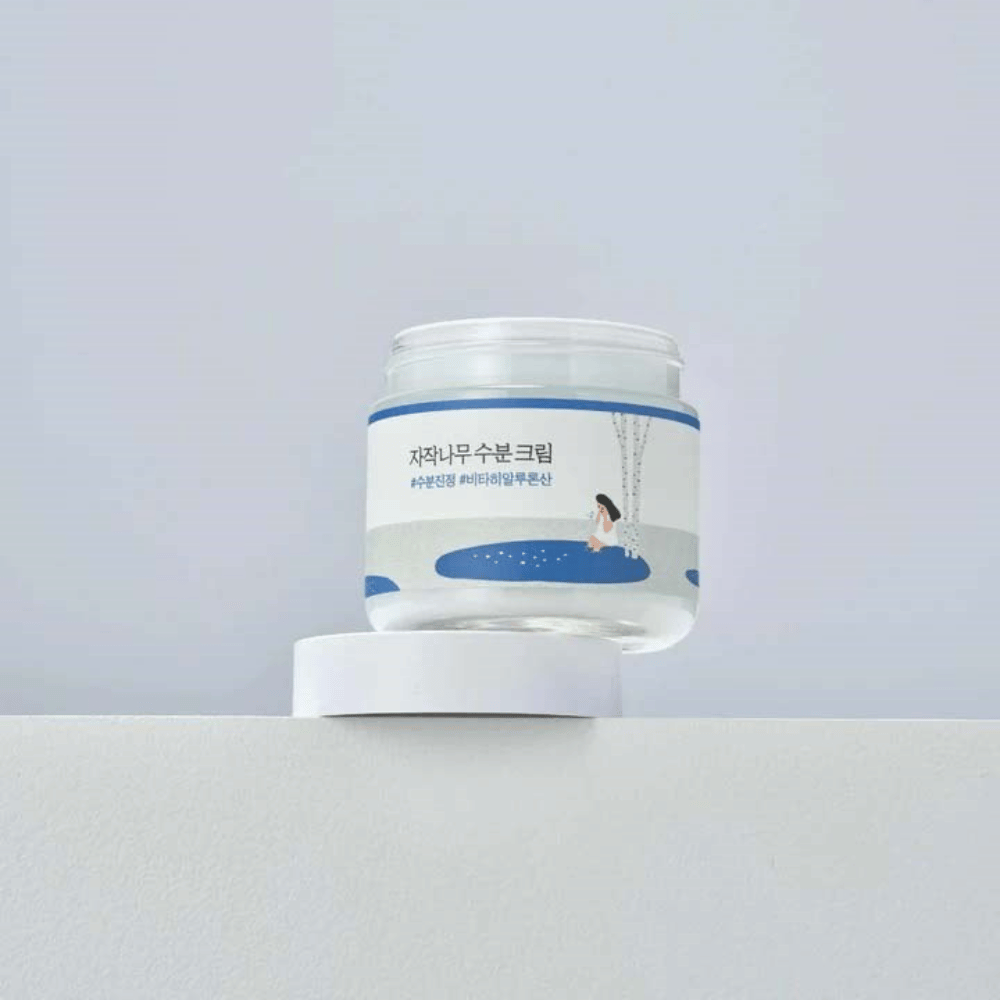 Round Lab Birch Juice Moisturizing Cream v priehľadnej nádobe s bielou etiketou a modrou ilustráciou ženy pri breze, stojaci na bielom povrchu proti svetlému pozadiu.
