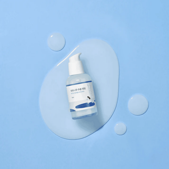 Round Lab Birch Juice Moisturizing Serum v priehľadnej pumpovacej fľaši umiestnenej na modrom pozadí s kvapkami a rozliatym sérom.