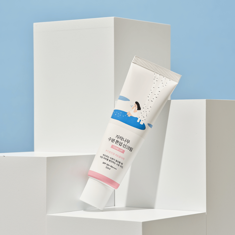 Tuba s Birch Juice Moisturizing Tone Up Sunscreen umiestnená na bielych kockách na svetlomodrom pozadí. Tuba je biela s ilustráciou ženy pri breze a vode. Text v kórejčine uvádza, že produkt má SPF 50+ PA++++ a obsahuje 50 ml.