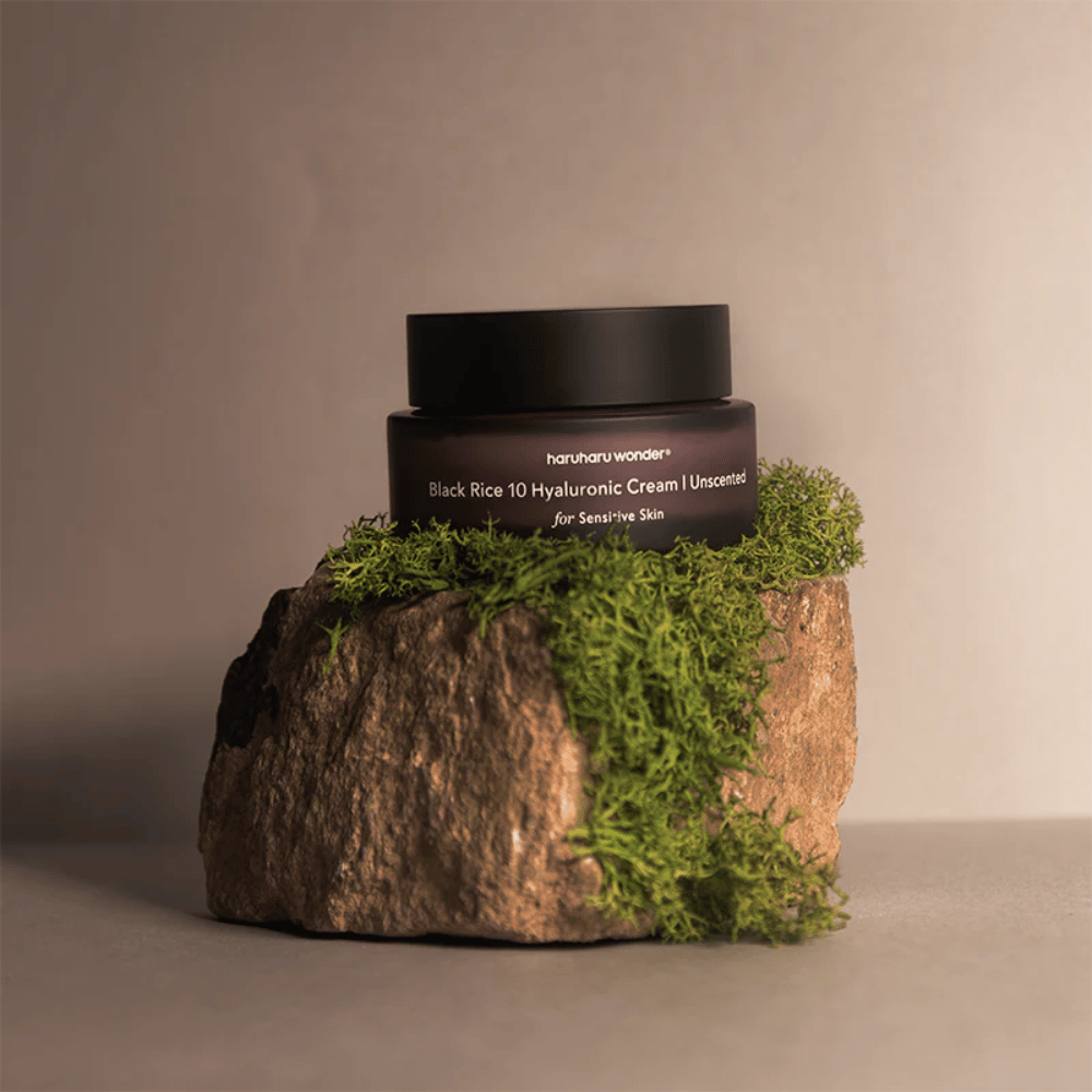 Obrázok zobrazuje nádobu s krémom na pleť označenú "haruharu wonder Black Rice 10 Hyaluronic Cream Unscented for Sensitive Skin". Nádoba je čierna a stojí na veľkom kameni pokrytom svetlozeleným machom, čo vytvára prirodzený a zemný pocit.