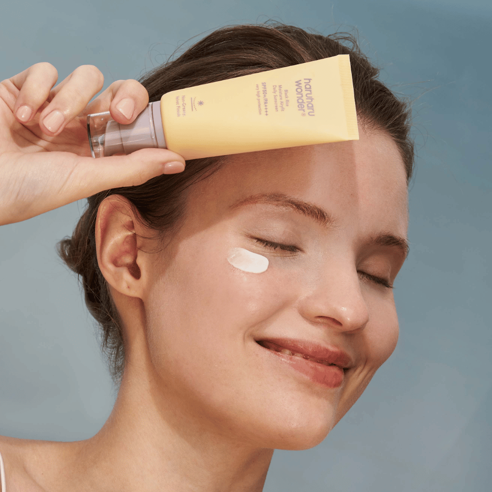 Usmievajúca sa žena aplikuje Black Rice Moisture Airyfit Daily Sunscreen SPF 50+ PA++++ od Haruharu Wonder na svoju tvár. Drží žltú tubu s opaľovacím krémom a má kvapku produktu na líci. Pozadie je svetlomodré.