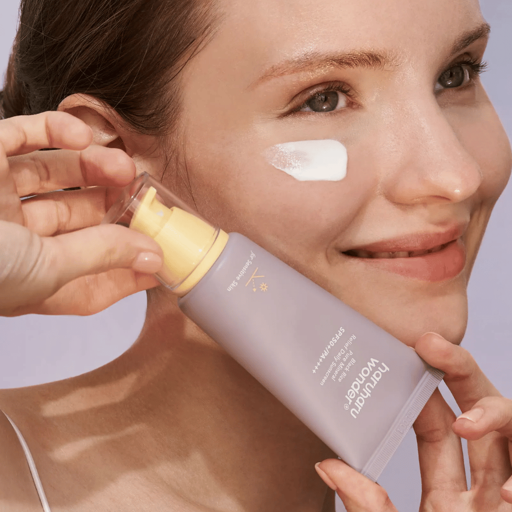 Usmievajúca sa žena nanáša Haruharu Wonder Black Rice Pure Mineral Relief Daily Sunscreen SPF 50+ PA++++ na svoju tvár. Drží fialovú tubu s opaľovacím krémom a má kvapku produktu na líci. Pozadie je svetlofialové.