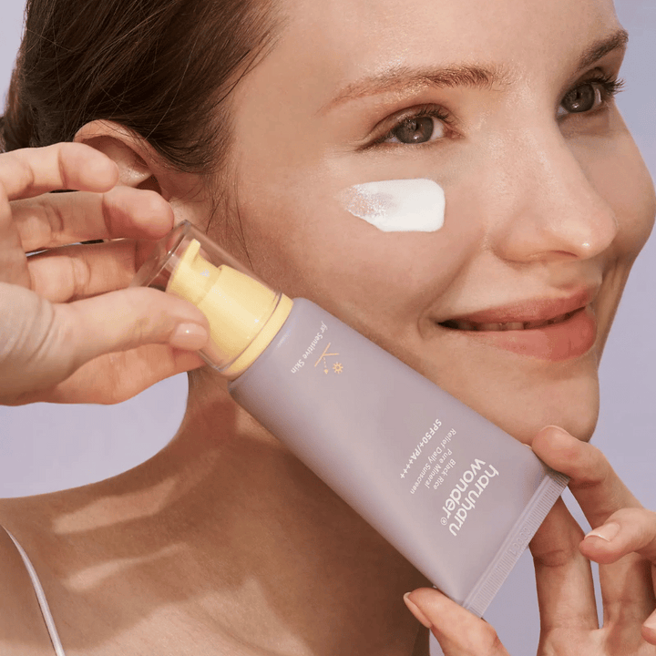 Usmievajúca sa žena nanáša Haruharu Wonder Black Rice Pure Mineral Relief Daily Sunscreen SPF 50+ PA++++ na svoju tvár. Drží fialovú tubu s opaľovacím krémom a má kvapku produktu na líci. Pozadie je svetlofialové.