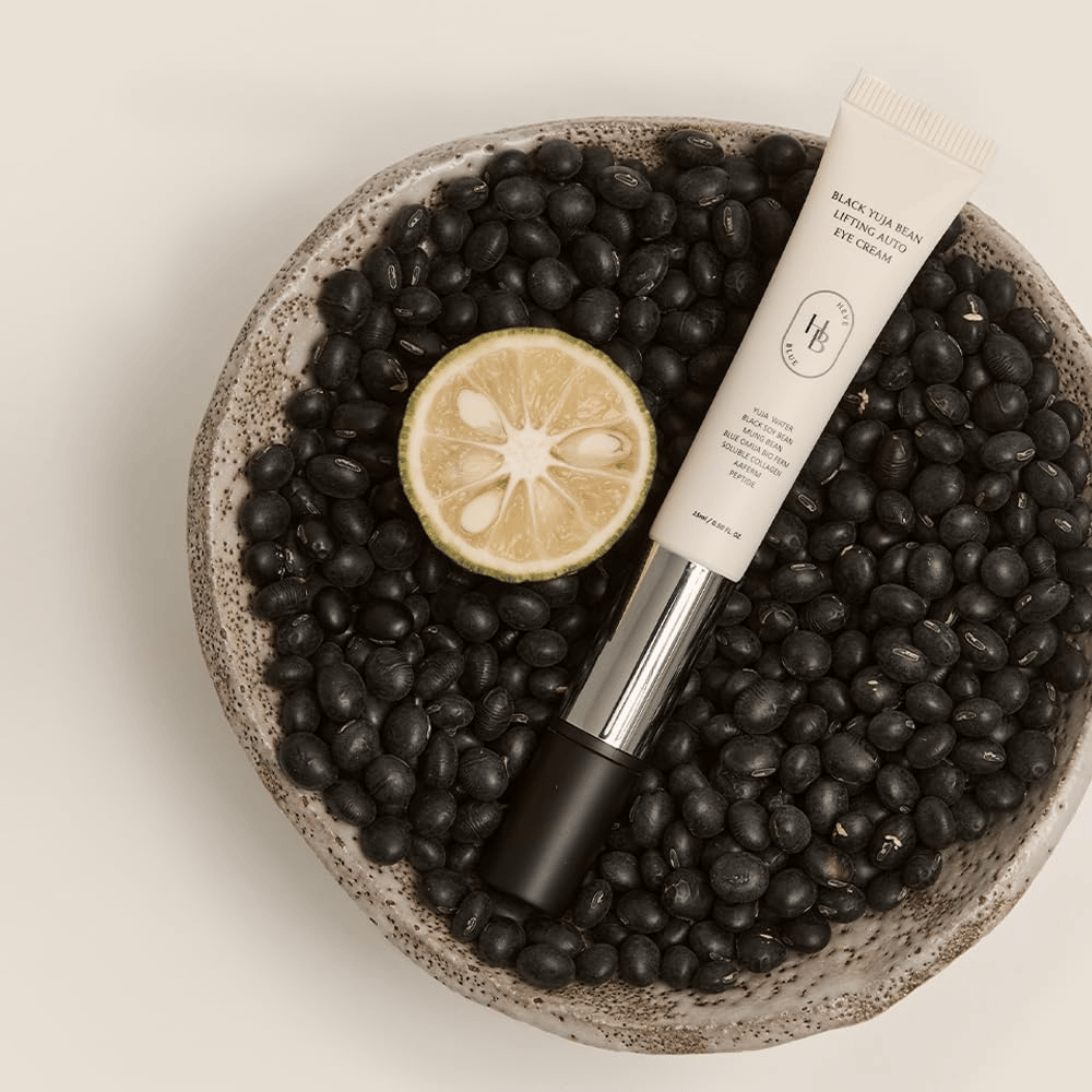 Black Yuja Bean Lifting Auto Eye Cream ležiaci v miske plnej čiernych fazúľ a polovičky citrusového plodu - vizuálna reprezentácia prírodných zložiek produktu.