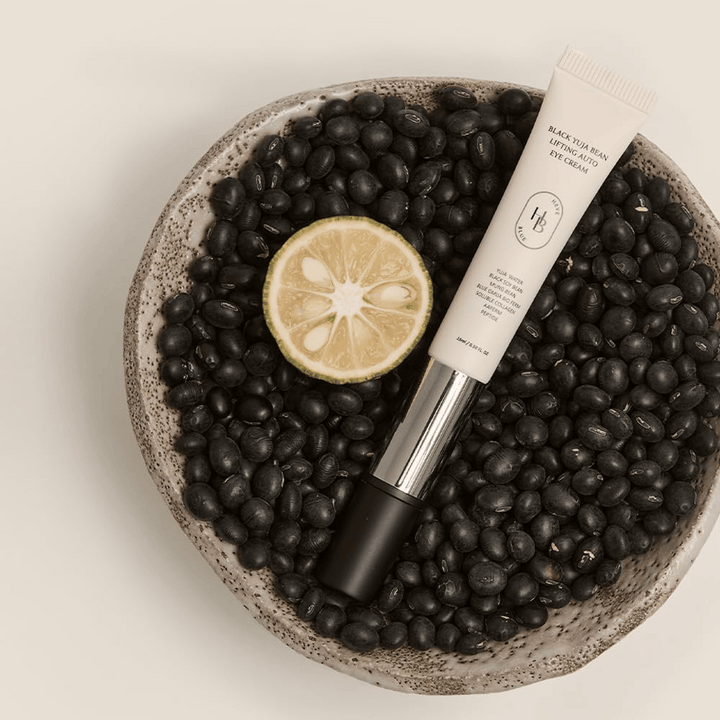 Black Yuja Bean Lifting Auto Eye Cream ležiaci v miske plnej čiernych fazúľ a polovičky citrusového plodu - vizuálna reprezentácia prírodných zložiek produktu.