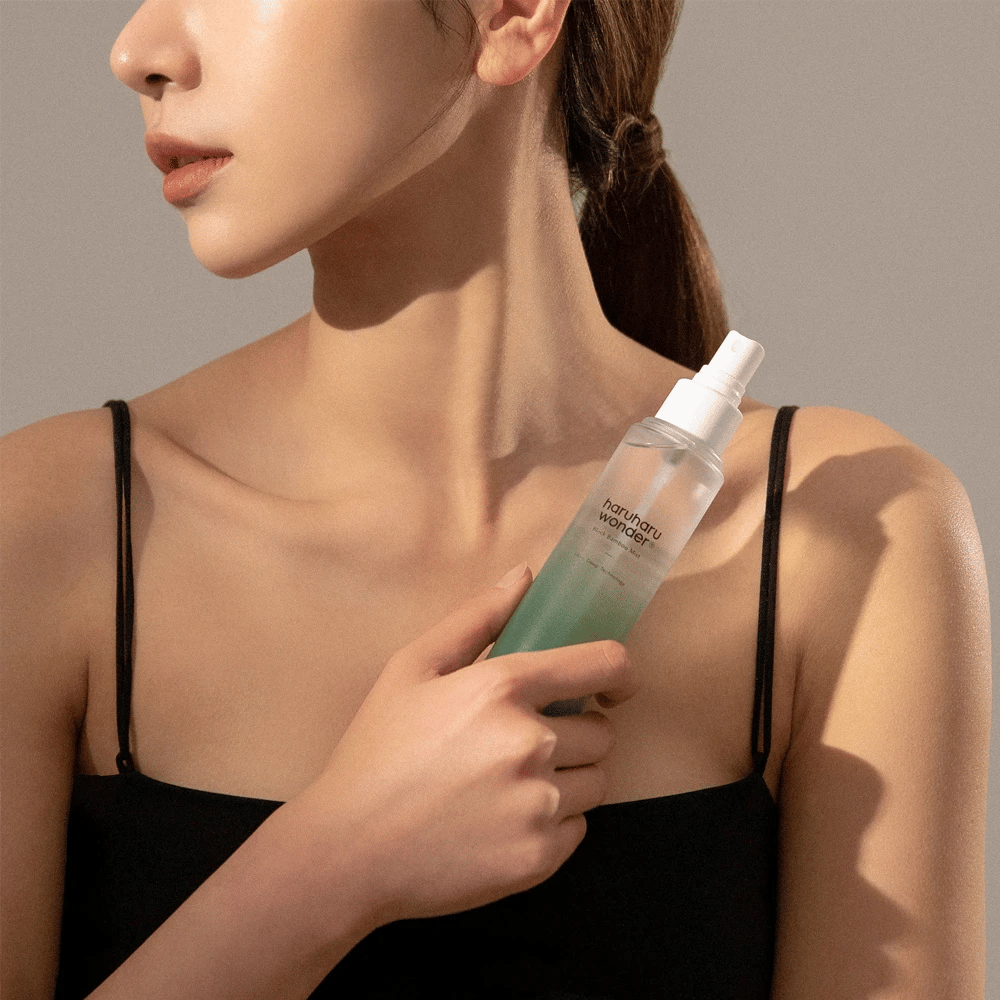 Obrázok HaruHaru Wonder Black Bamboo Mist držiaci žena. Produkt je v priehľadnej sprejovej fľaši so zeleným odtieňom a bielou pumpičkou. Žena má uvoľnený vzhľad, nosí čierny top a sústredí sa na použitie hmly na dodanie hydratácie a lesku pokožke. Fľaša je súčasťou starostlivosti o pleť.