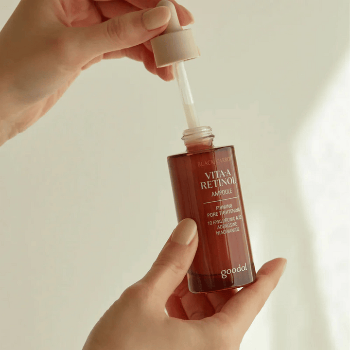 Ruka drží fľašu s Goodal Black Carrot Vita-A Retinol Firming Ampoule. Etiketa na fľaši zobrazuje názov produktu a jeho vlastnosti, vrátane "Firming Pore Tightening" a ingrediencií ako 10% kyseliny hyalurónovej, adenosín a niacínamid. Pipeta je v béžovej farbe.